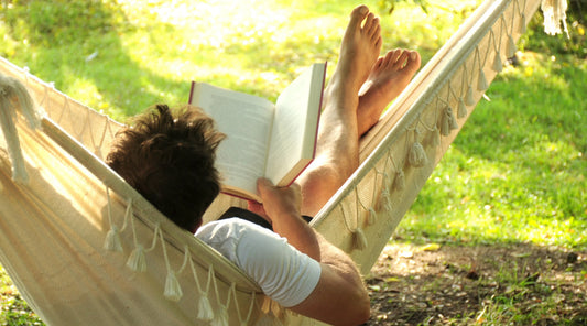 Person relaxing in a hammock while reading a book outdoors in warm sunlight, illustrating the importance of rest for wellbeing, relaxation and recovery as part of a healthy lifestyle.