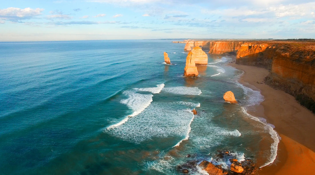 Scenic aerial view of the Twelve Apostles along the Great Ocean Road, with waves crashing onto the beach and the rugged cliffs illuminated by sunlight