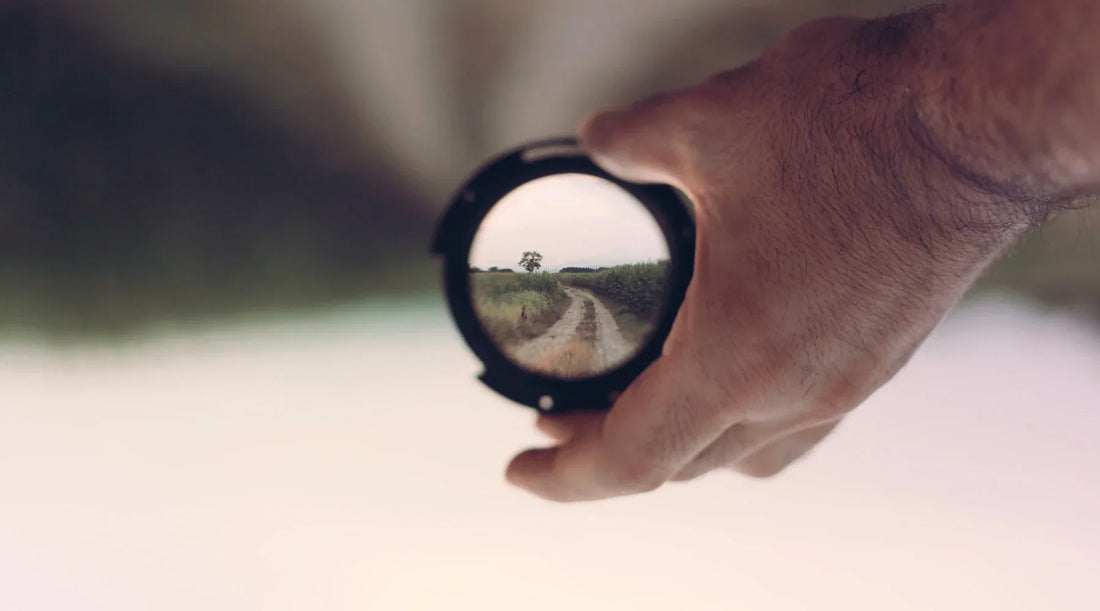 Close-up of a hand holding a lens filter focused on a dirt path and lone tree, symbolising clarity, perspective, and staying focused on long-term wellness goals.