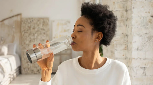 A woman wearing a white top drinks water from a clear glass bottle with a crystal inside, standing in a bright, softly lit room with a white brick wall in the background.