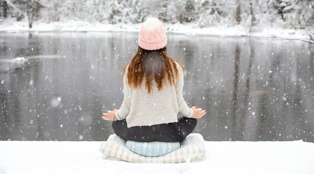 Woman practising mindfulness meditation outdoors during winter, sitting beside a snow-covered lake surrounded by falling snow, representing winter wellbeing, seasonal self-care, stress management, relaxation, and holistic wellness.
