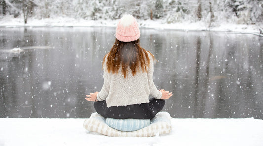 Woman practising mindfulness meditation outdoors during winter, sitting beside a snow-covered lake surrounded by falling snow, representing winter wellbeing, seasonal self-care, stress management, relaxation, and holistic wellness.