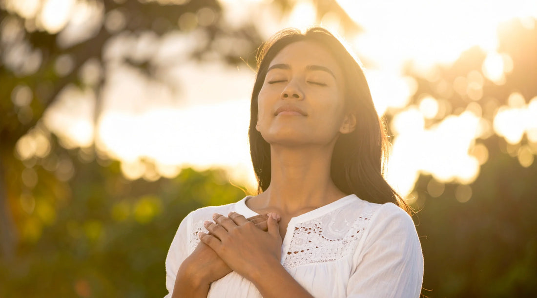Woman standing outdoors with eyes closed and hands on chest in sunlight, representing calm breathing, nervous system regulation, emotional balance, and holistic wellness.
