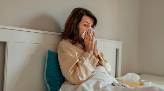 Woman sitting up in bed with a tissue, appearing unwell and blowing her nose, with tea and medicine on the bedside table.