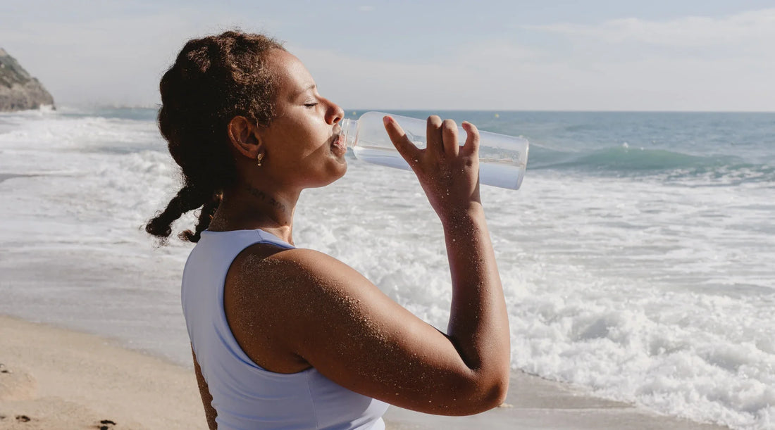 Woman drinking water on a sunny beach, representing hydration, wellness, and springtime gut health in Australia.