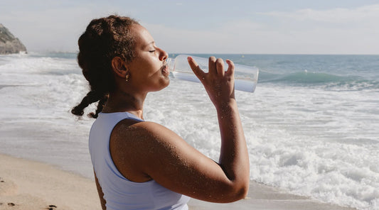 Woman drinking water on a sunny beach, representing hydration, wellness, and springtime gut health in Australia.