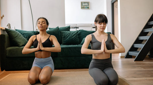 Two women practising yoga indoors with hands in prayer position, representing mindfulness, nervous system regulation, stress support, and gentle daily rituals that help create sustainable focus and balanced energy.
