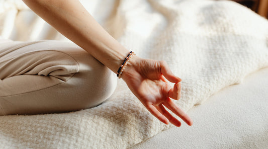 A close-up of a person sitting cross-legged on a soft beige blanket, resting their hand in a gentle meditation mudra, symbolising calm, relaxation, and slow-season wellness.
