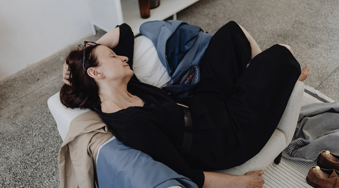 An exhausted woman lying curled up on a chair with her eyes closed, surrounded by clothes and shoes on the floor — illustrating burnout, holiday fatigue, and the physical impact of stress