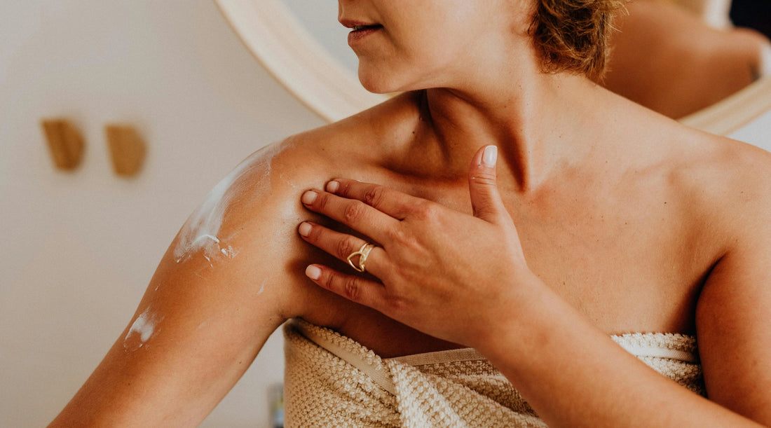 A woman wrapped in a towel applying moisturiser to her shoulder, showing a close-up self-care moment focused on skin hydration and relaxing daily wellness rituals.