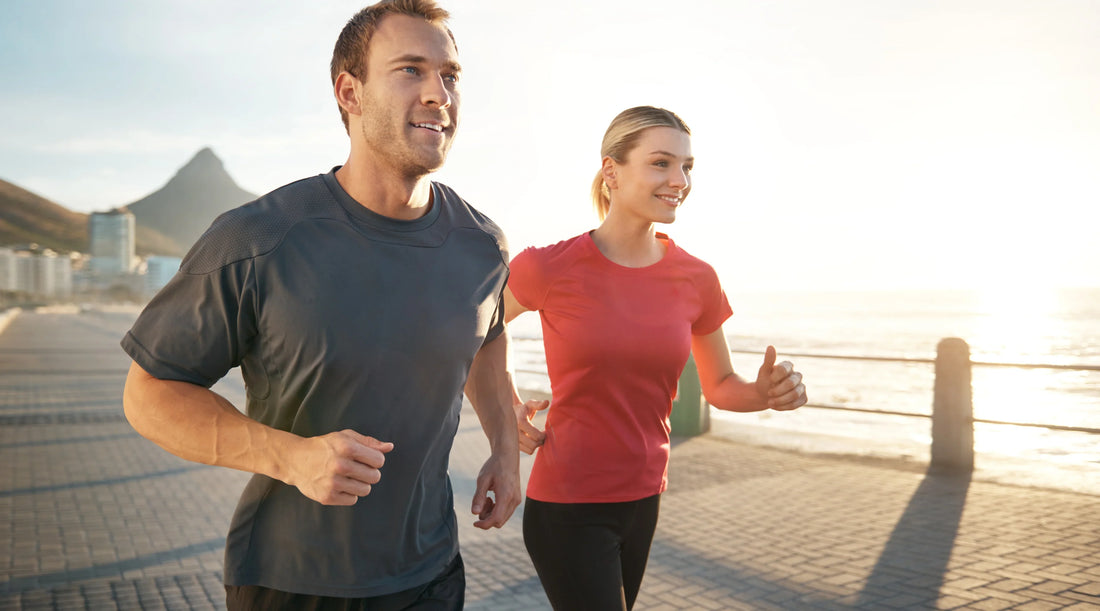 A man and woman jogging outdoors along a seaside path at sunrise, smiling and enjoying an energising workout representing active lifestyle, movement, and natural stress relief