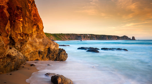 Golden coastal cliffs meeting a calm turquoise ocean at sunset, with soft waves washing over sandy shore and rock formations, reflecting a sense of balance, resilience, and nature-inspired wellbeing.