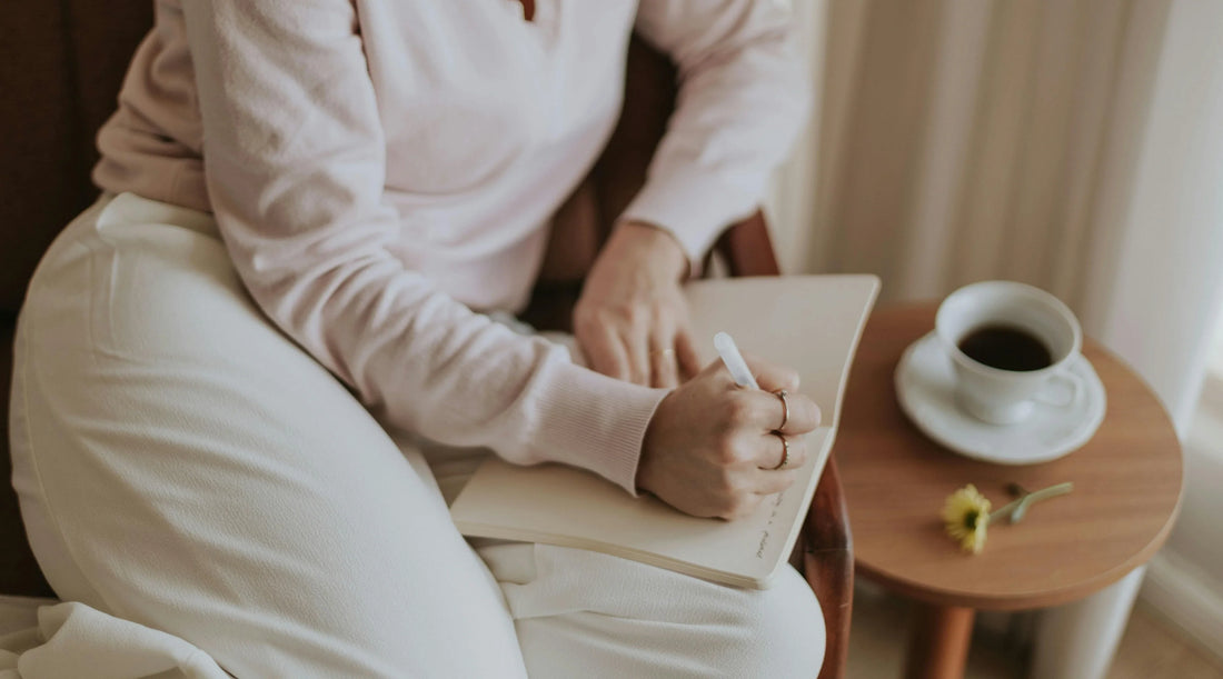 Person journaling in a notebook beside coffee in a calm home setting, representing mindful reflection, intentional habits, emotional clarity, slow living, and holistic wellbeing routine