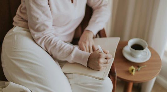 Person journaling in a notebook beside coffee in a calm home setting, representing mindful reflection, intentional habits, emotional clarity, slow living, and holistic wellbeing routine