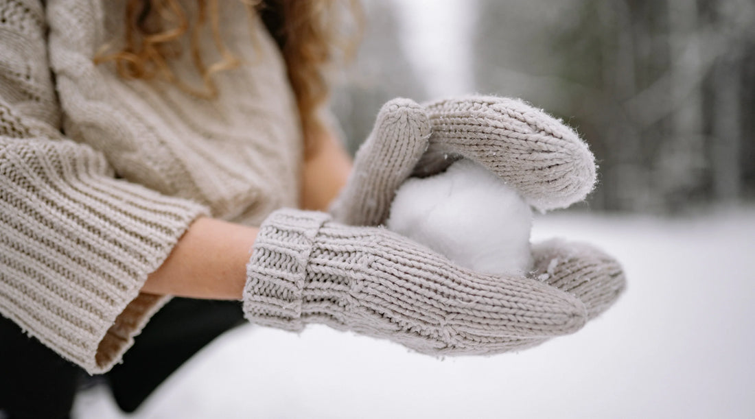 A person wearing a cosy knitted jumper and gloves holding a snowball outdoors in winter. The image represents seasonal wellness, staying warm, mindful living, winter self-care, and healthy daily habits during the colder months.