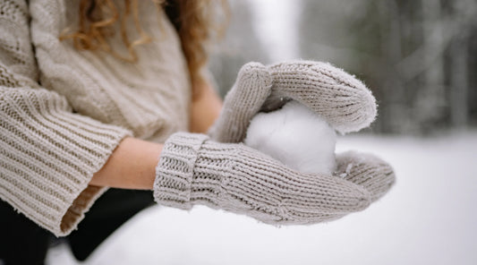 A person wearing a cosy knitted jumper and gloves holding a snowball outdoors in winter. The image represents seasonal wellness, staying warm, mindful living, winter self-care, and healthy daily habits during the colder months.