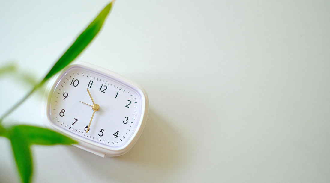 Minimal white clock beside green leaves symbolising slow living, stress management, rest, recovery, balanced routines, mental wellbeing, sleep support, and sustainable wellness habits.