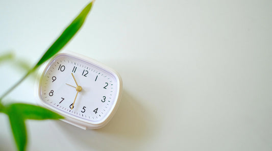Minimal white clock beside green leaves symbolising slow living, stress management, rest, recovery, balanced routines, mental wellbeing, sleep support, and sustainable wellness habits.